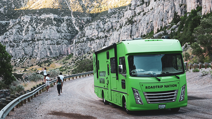 The roadtrippers explore a picturesque gorge with the green RV parked in the foreground.